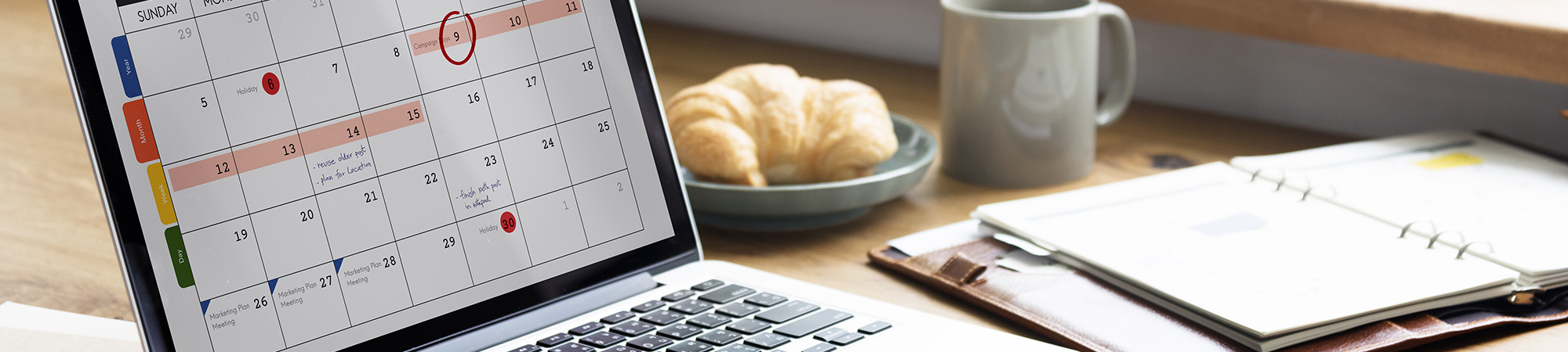 Online calendar displayed on laptop on desk with coffee, muffin, and planner