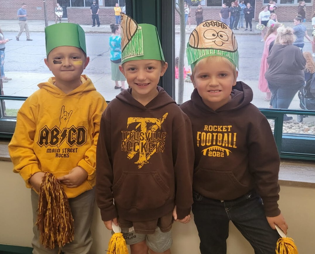 Three happy early childhood learning center students wearing green paper hats and brown and gold attire